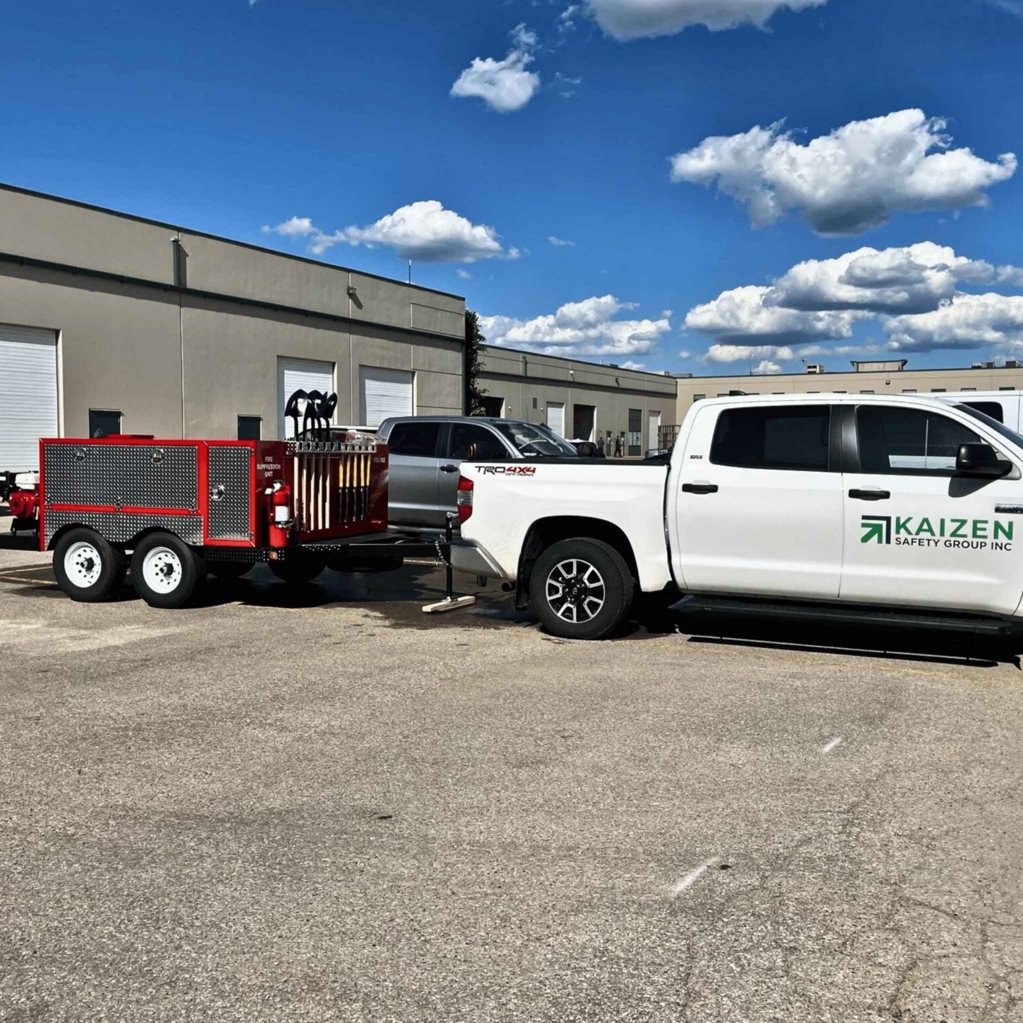 White truck with 'Kaizen Safety Group Inc' branding towing a red trailer in an industrial area.